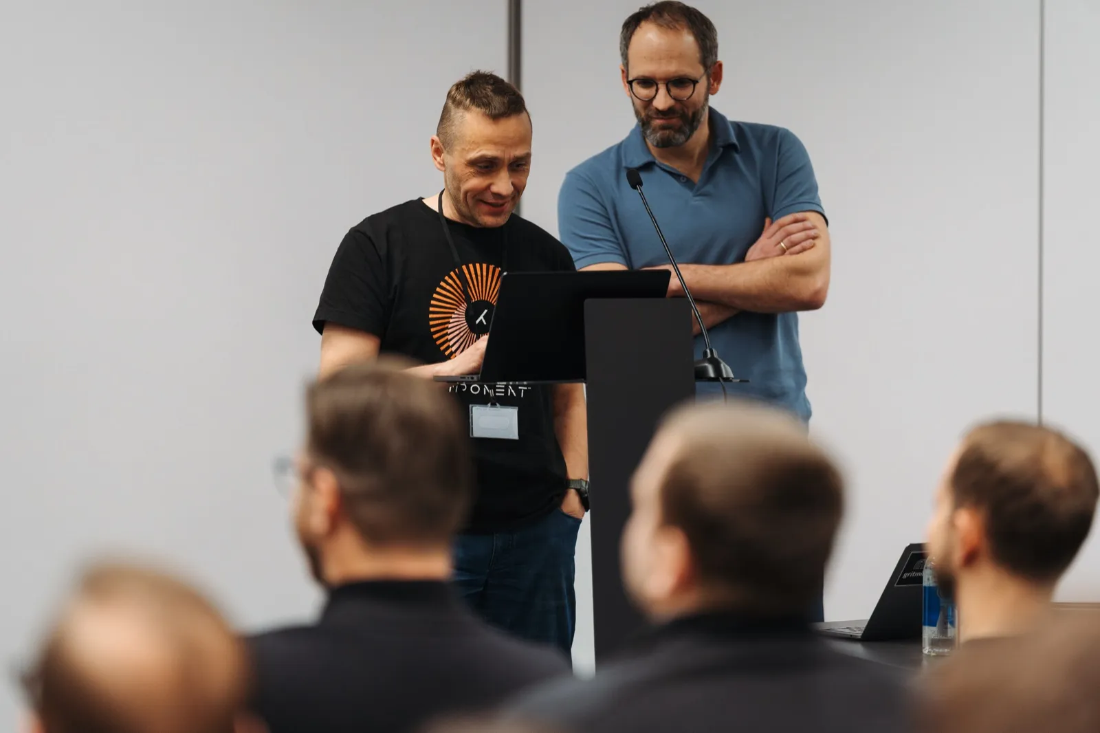 Two presenters at a lectern reviewing a laptop before a talk